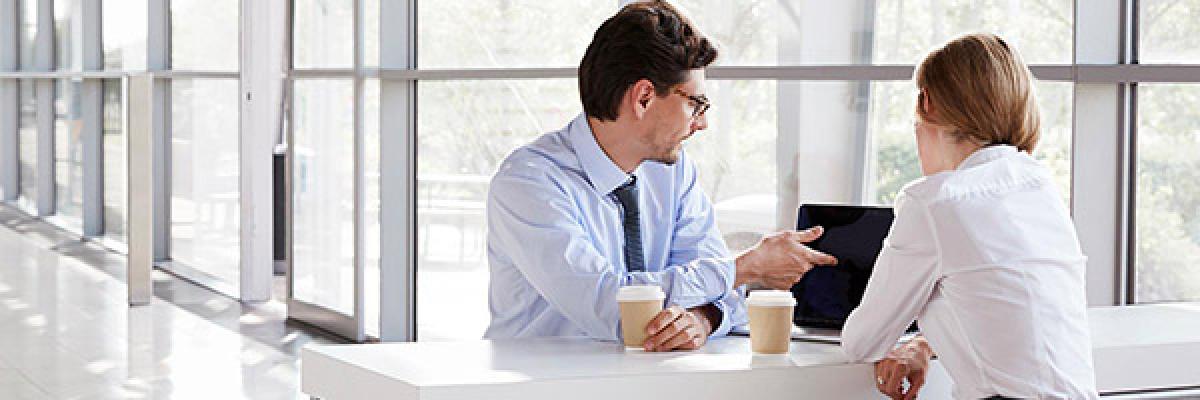 Financiación internacional - Reunión de negocios entre un hombre y una mujer de negocios con camisa, frente a un portátil hablando y tomando café Financiación internacional - Reunión de negocios entre un hombre y una mujer de negocios con camisa, frente a un portátil hablando y tomando café